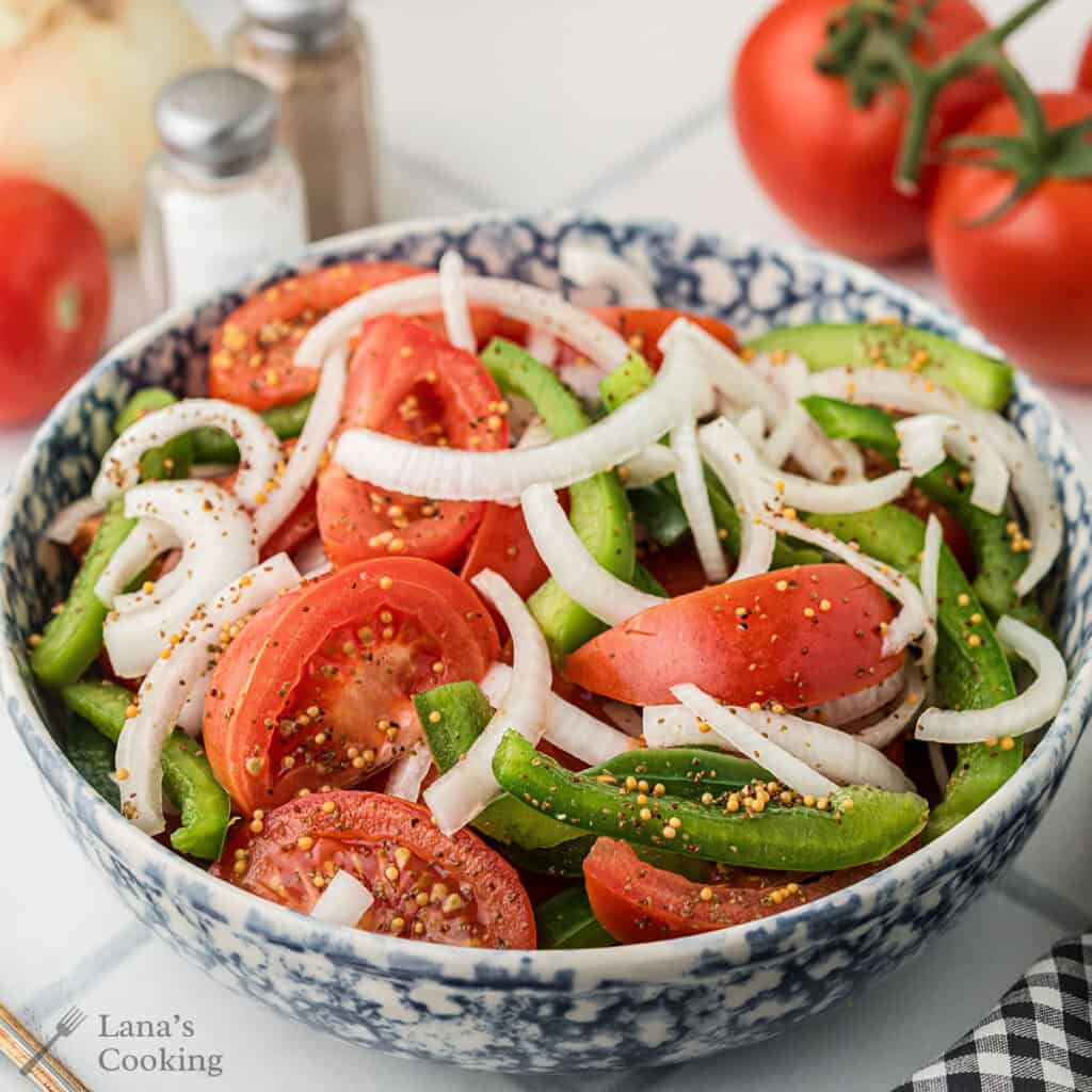 A bowl of sliced tomatoes, green peppers, and onions sprinkled with seasoning.