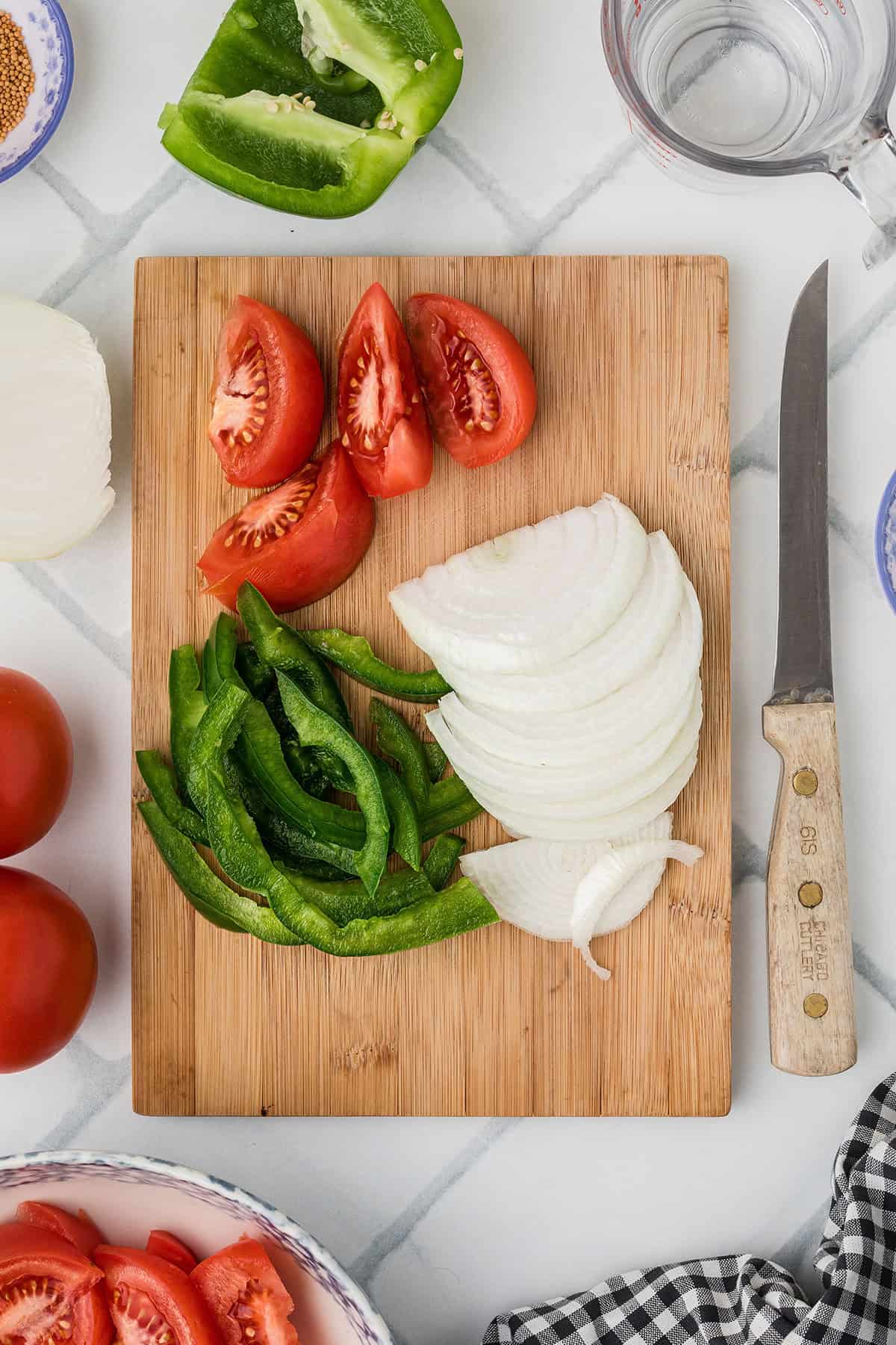 Sliced tomatoes, green bell pepper, and onion on a wooden cutting board with a knife and veggies nearby.