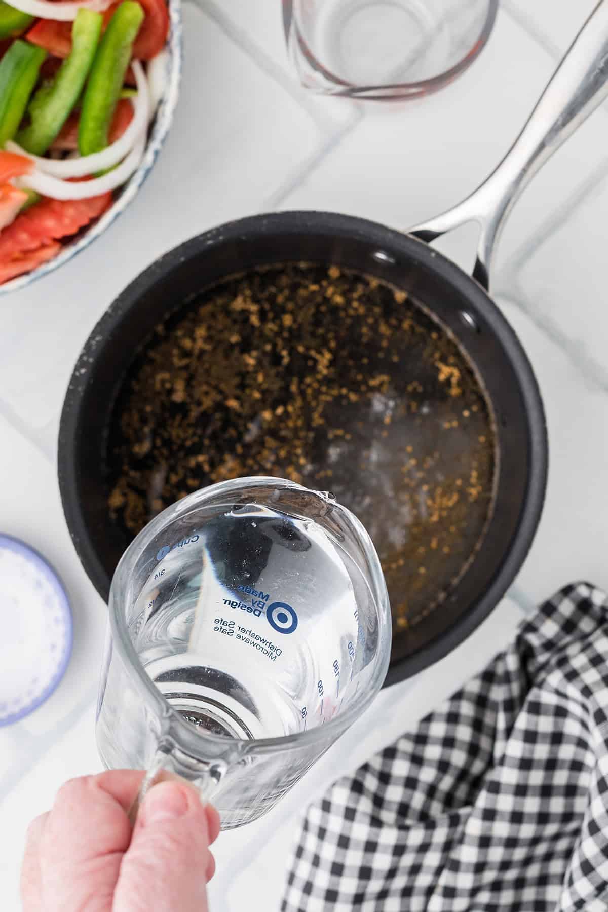 A hand pours water from a measuring cup into a black saucepan on a white counter near vegetables.