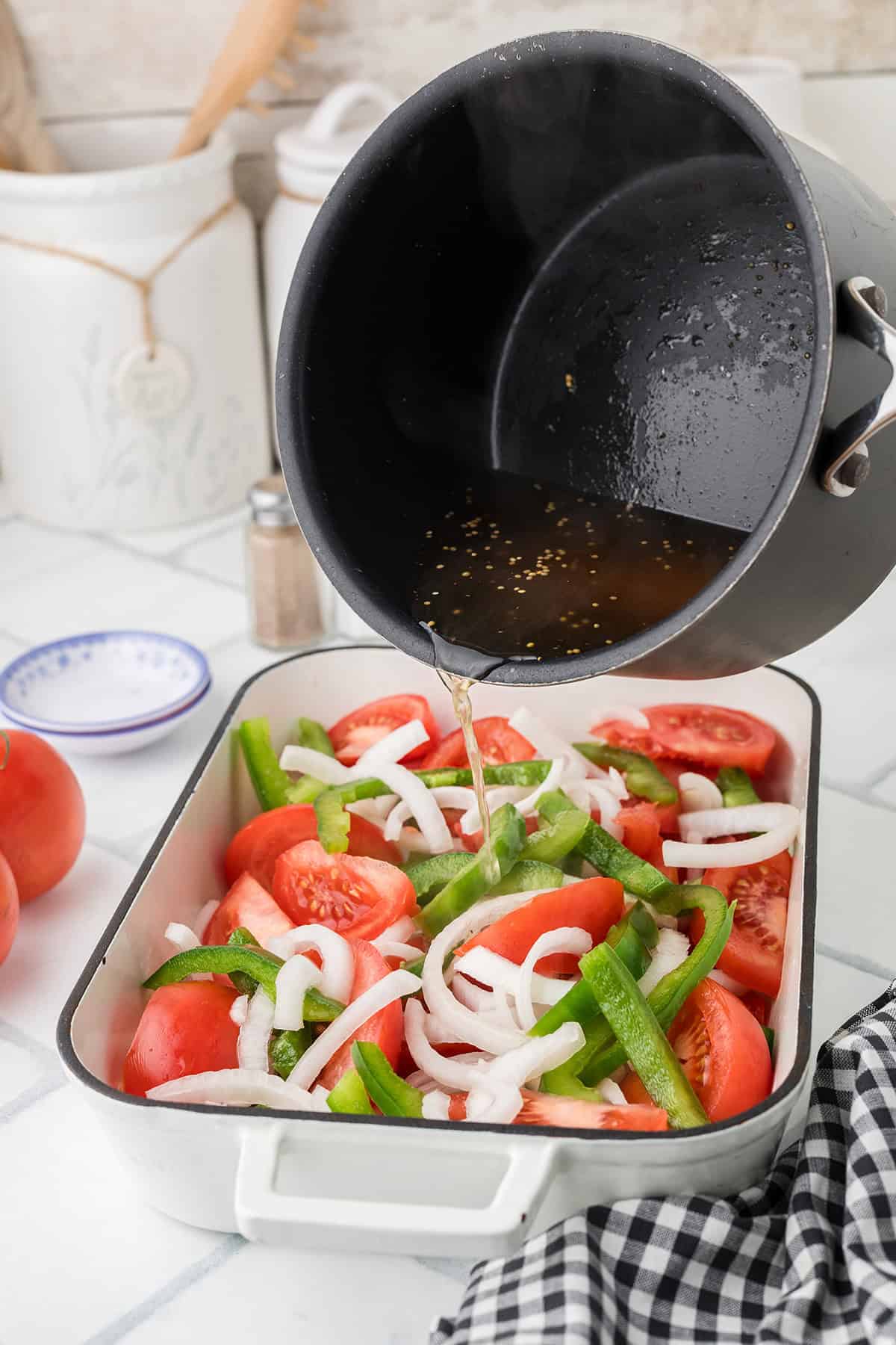 Broth is being poured from a pot over sliced tomatoes, onions, and green peppers in a baking dish.