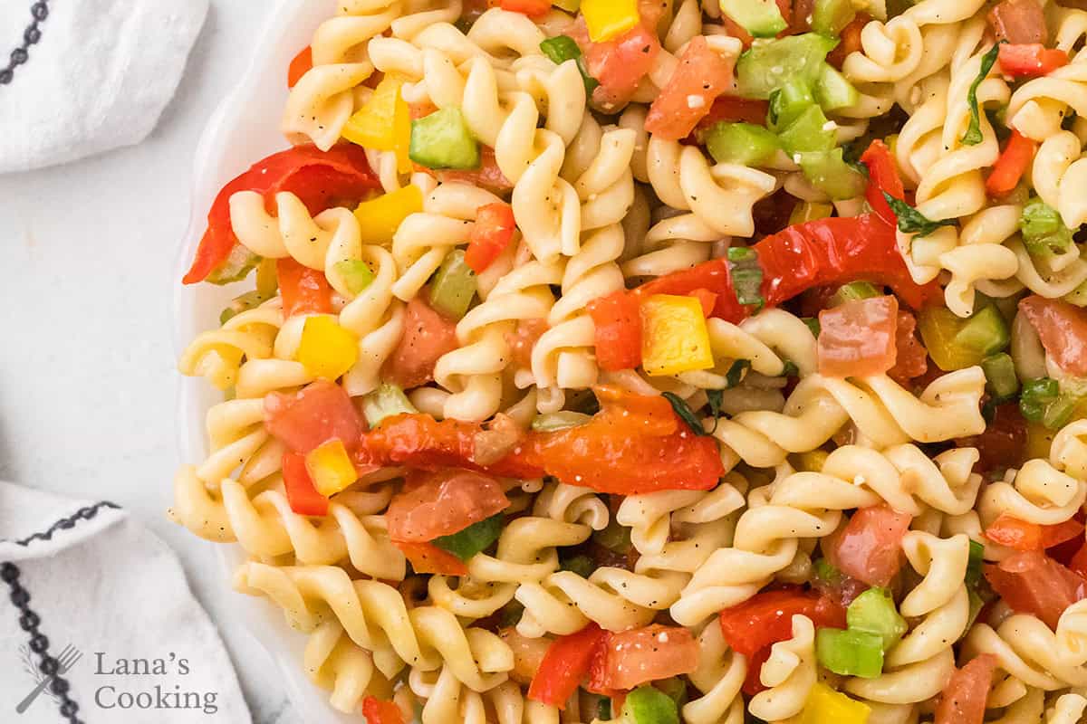 A close-up of pasta salad with rotini, diced tomatoes, bell peppers, and green vegetables on a white plate.