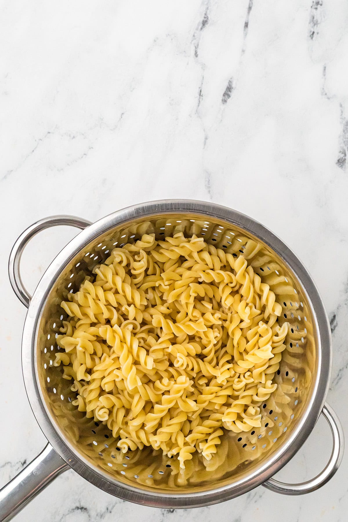 Cooked fusilli pasta draining in a stainless steel colander on a white marble countertop.