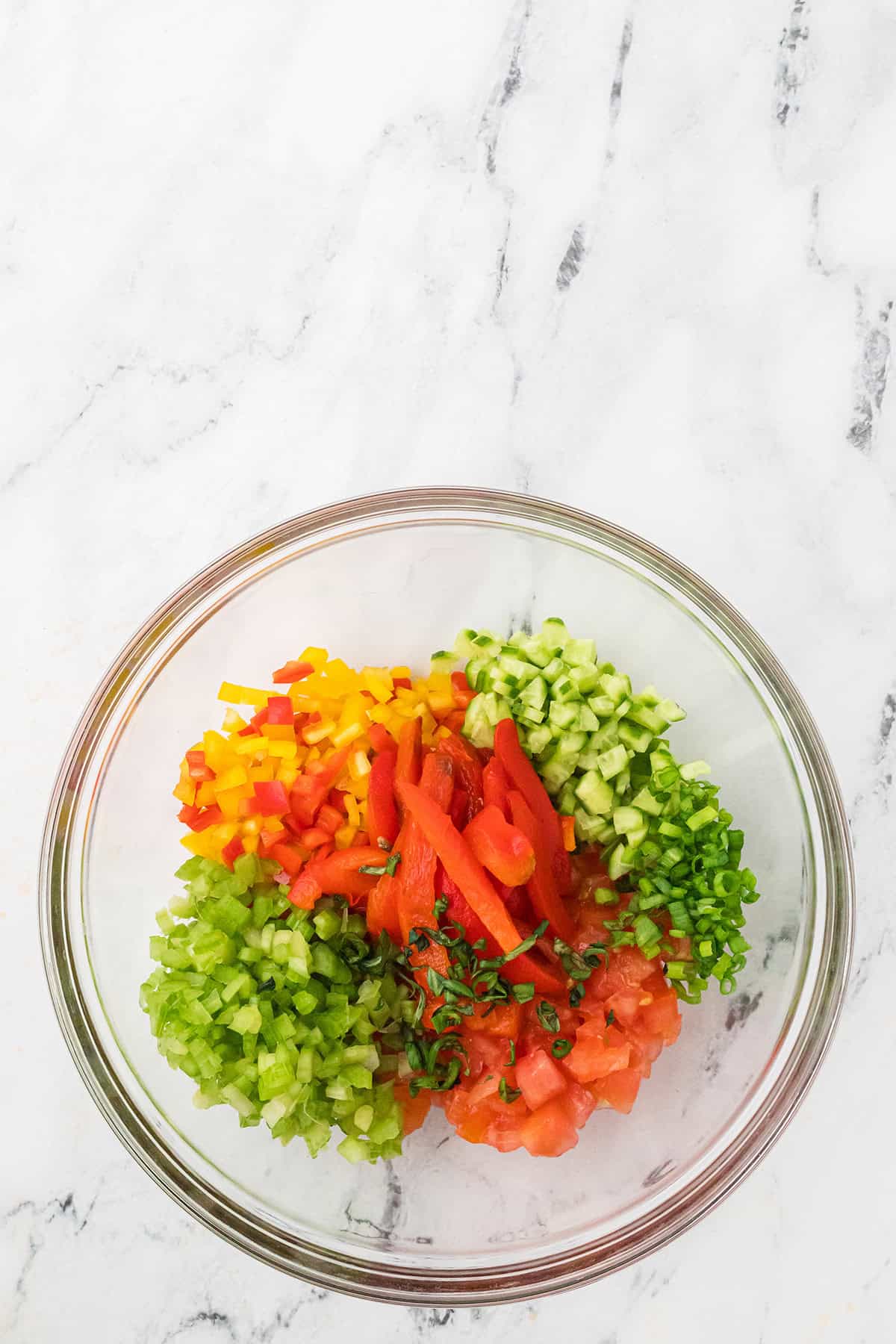 Chopped bell peppers, cucumber, tomato, and herbs arranged in a clear glass bowl on a marble surface.