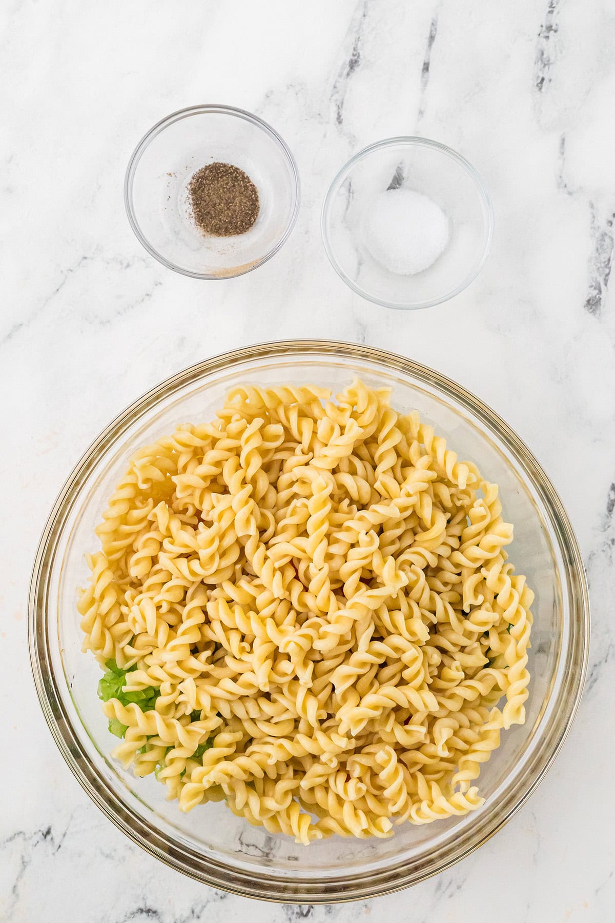 A glass bowl of cooked rotini pasta with two small bowls of salt and pepper on a marble surface.
