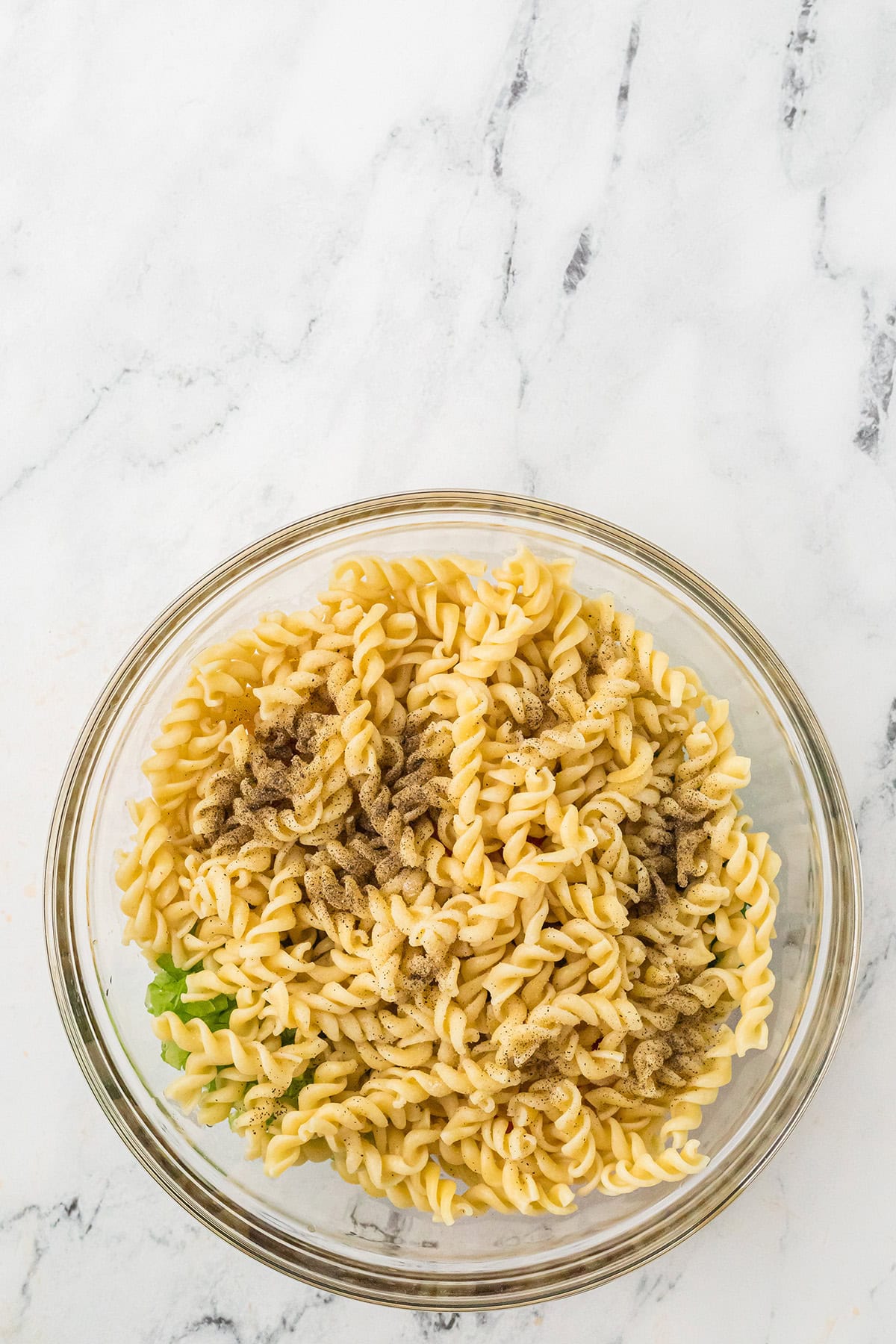 A glass bowl of cooked rotini pasta mixed with ground black pepper on a marble surface.
