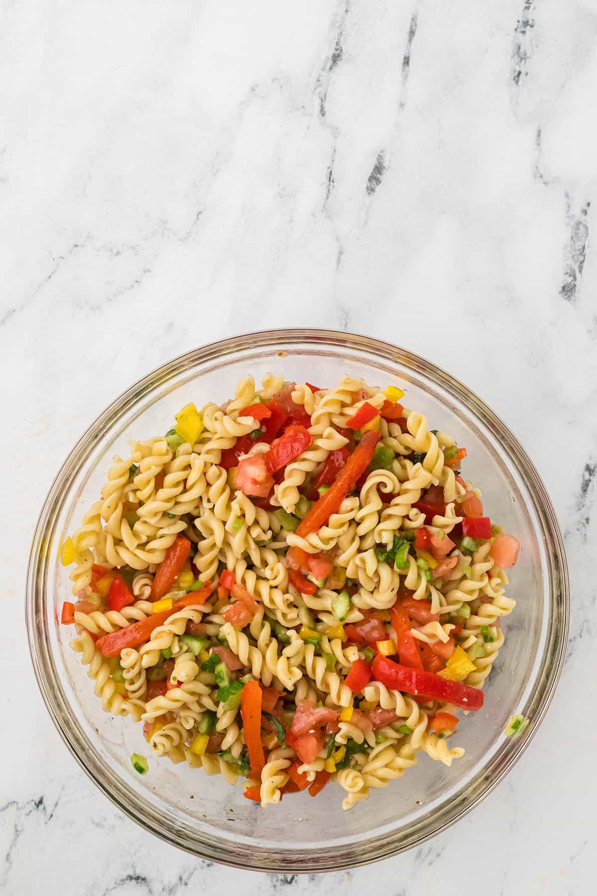 Glass bowl of rotini pasta salad with chopped tomatoes, bell peppers, and herbs on a marble surface.