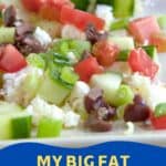 A close-up of a Greek appetizer with chopped tomatoes, cucumbers, olives, feta cheese, and green onions on a plate, with a pepperoncini in the background.