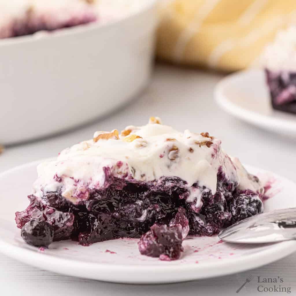 A slice of blueberry pineapple congealed salad with whipped topping and nuts on a white plate, with a spoon beside it.