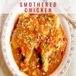 An oval plate with Creole-style smothered chicken in a thick, seasoned sauce, garnished with chopped green onions; fork and knife are placed beside the plate.