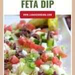 Close-up of a layered feta dip with cucumbers, tomatoes, olives, and green onions on a white plate.