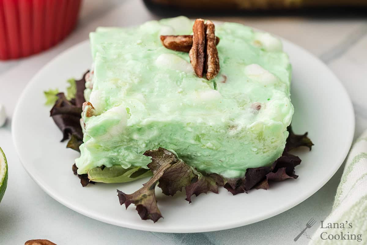 A slice of green gelatin salad with pecans and marshmallows, served on lettuce on a white plate.