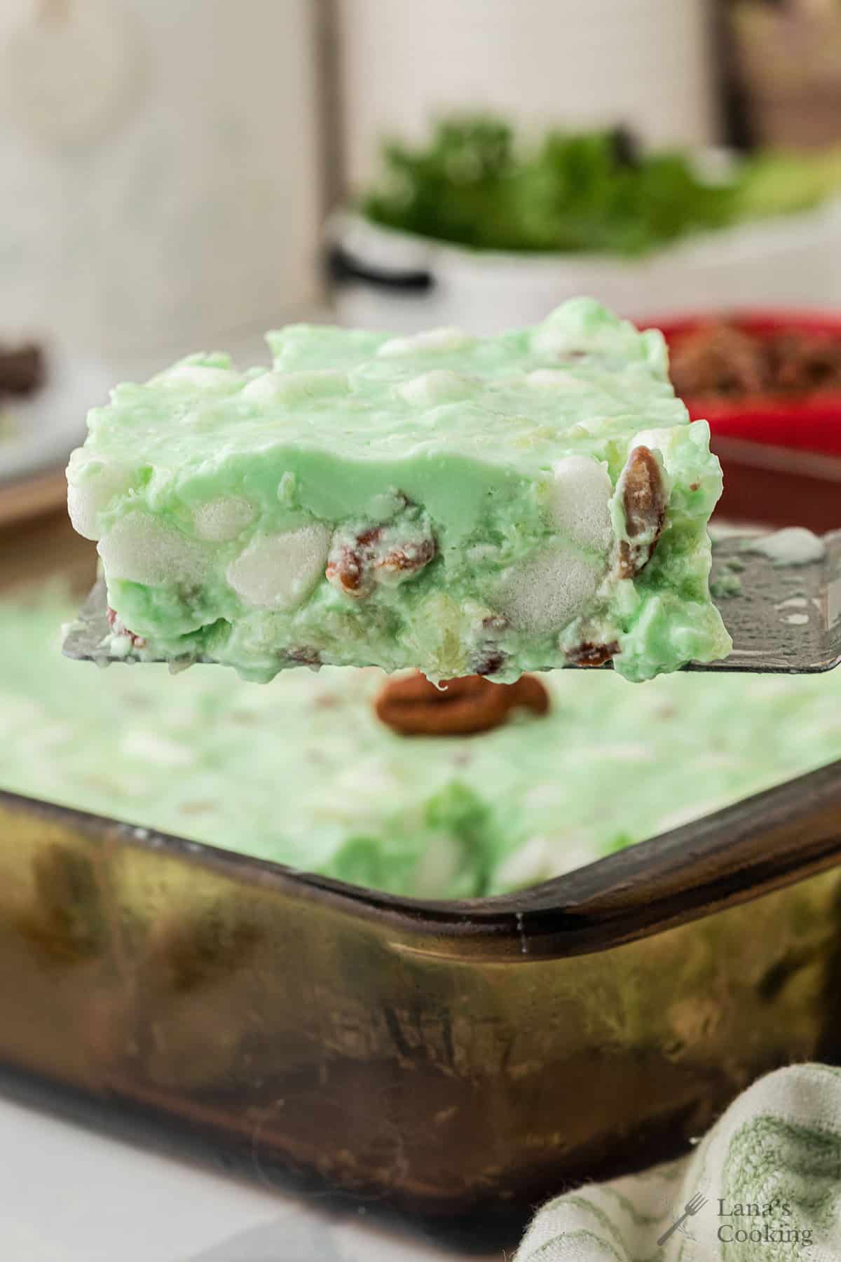 A creamy green jello salad with marshmallows and nuts being lifted from a glass baking dish.