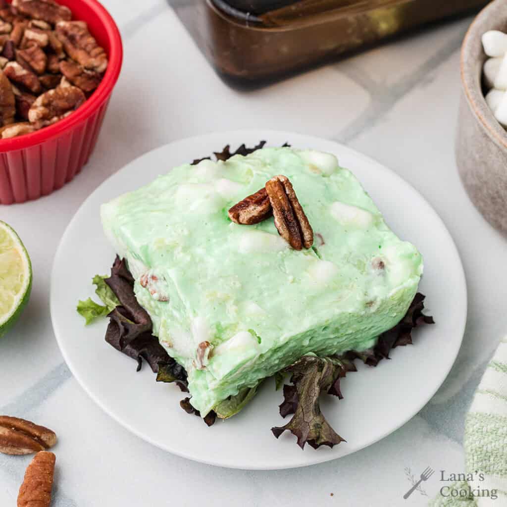 A slice of green congealed salad with pecans on top, served on lettuce on a white plate.