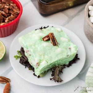 A slice of green congealed salad with pecans on top, served on lettuce on a white plate.