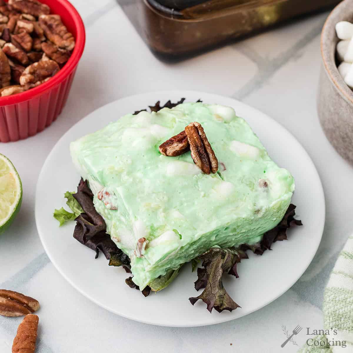 A slice of green congealed salad with pecans on top, served on lettuce on a white plate.