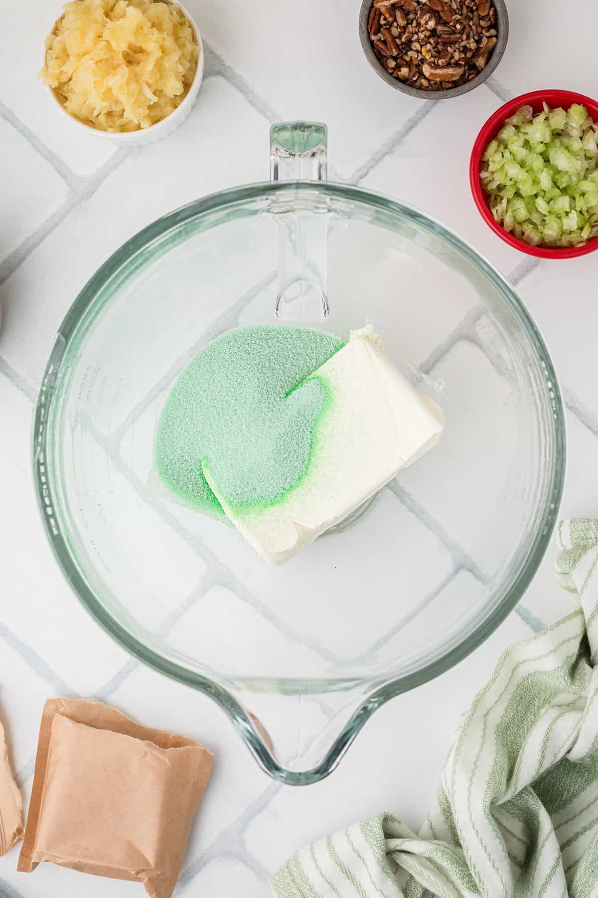 Glass bowl with green gelatin powder and cream cheese, surrounded by bowls of pineapple, pecans, and celery.