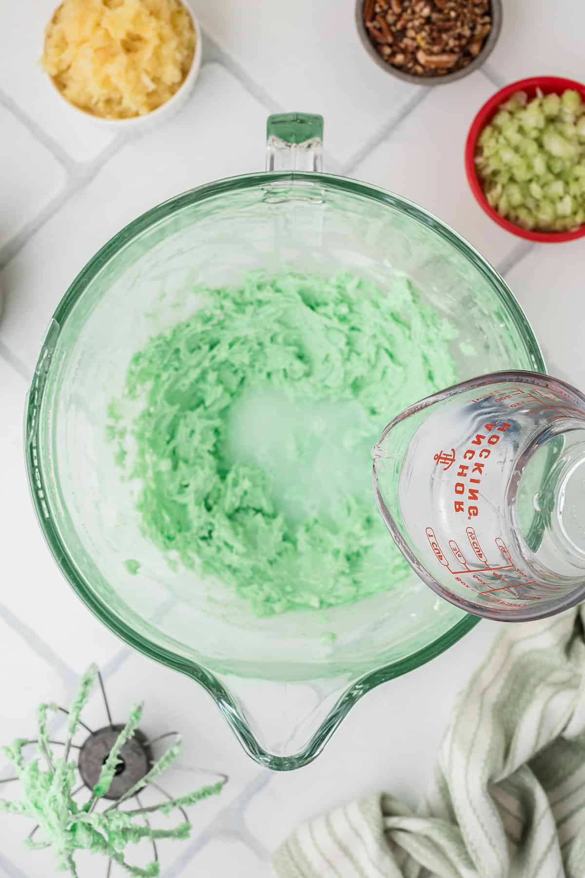 A glass mixing bowl with light green whipped mixture, a measuring cup of liquid, and small bowls of ingredients nearby.