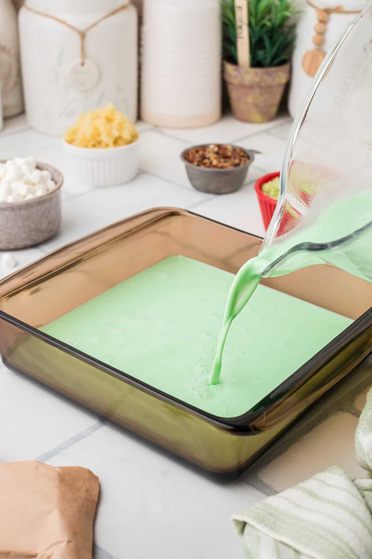 Green liquid is being poured from a glass pitcher into a square brown glass baking dish on a kitchen counter.