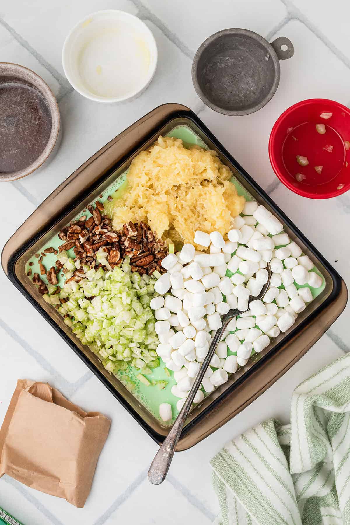 Square baking dish with marshmallows, pineapple, chopped celery, pecans, and a mixing spoon inside.