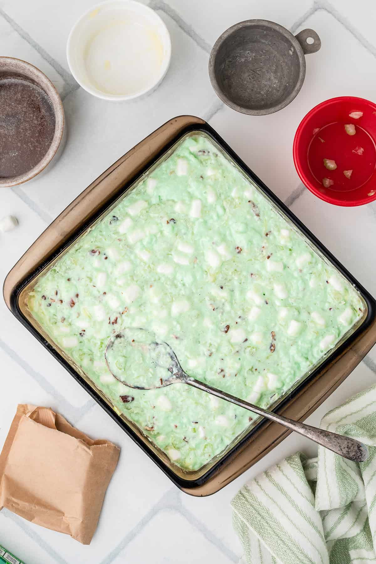 A square pan of green jello salad with marshmallows sits on a tile surface, surrounded by empty bowls.