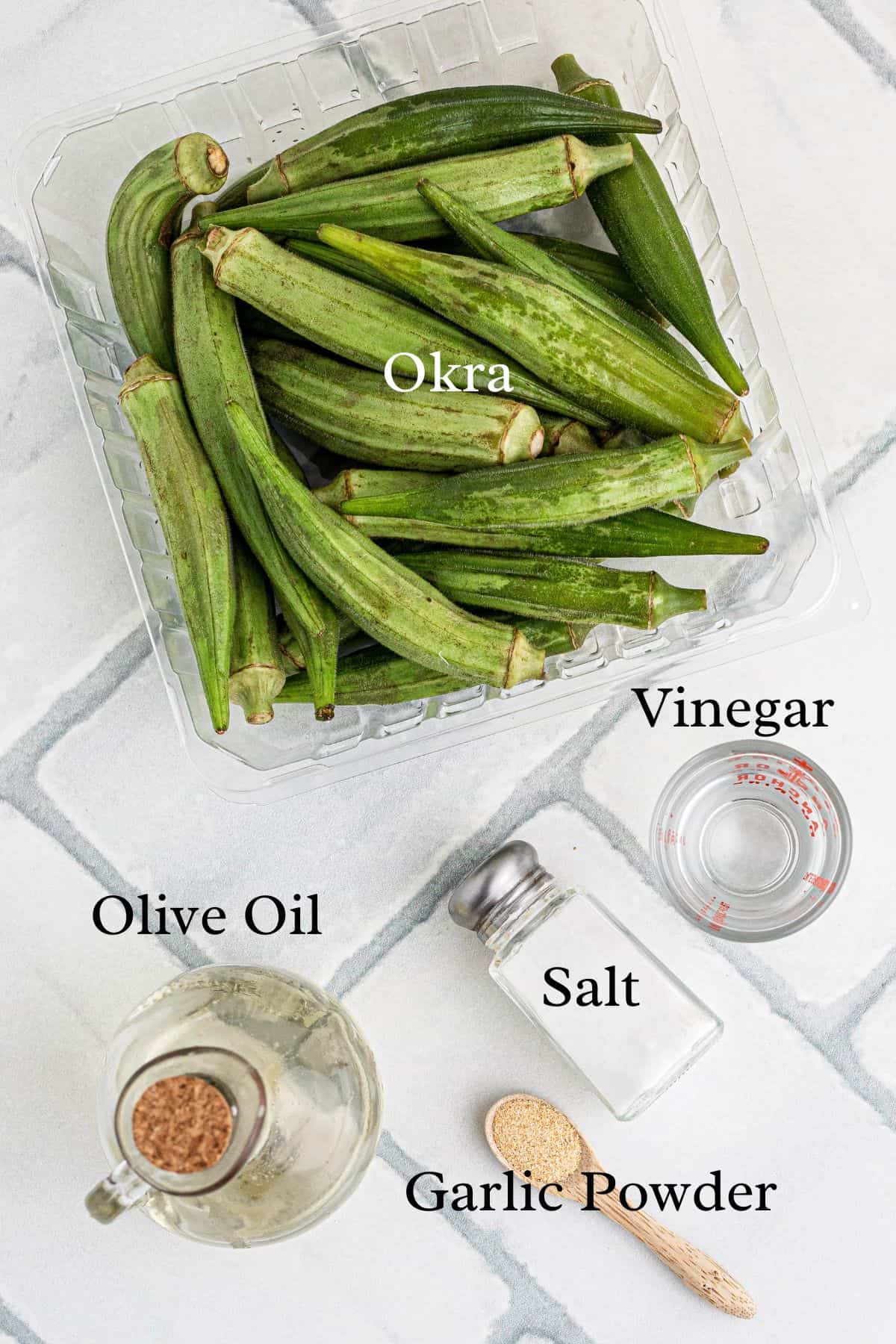 Plastic container of fresh okra, with olive oil, vinegar, salt, and garlic powder arranged on a tiled surface.