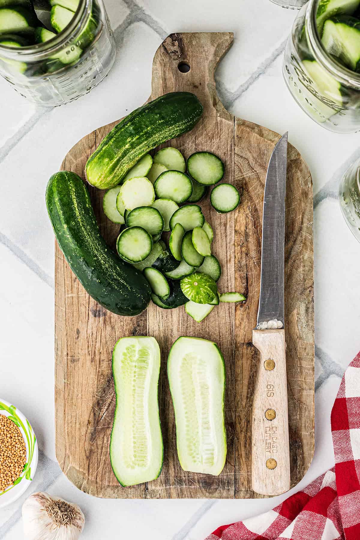 Sliced and whole cucumbers on a wooden cutting board with a knife, surrounded by glass jars and a red-checkered cloth.