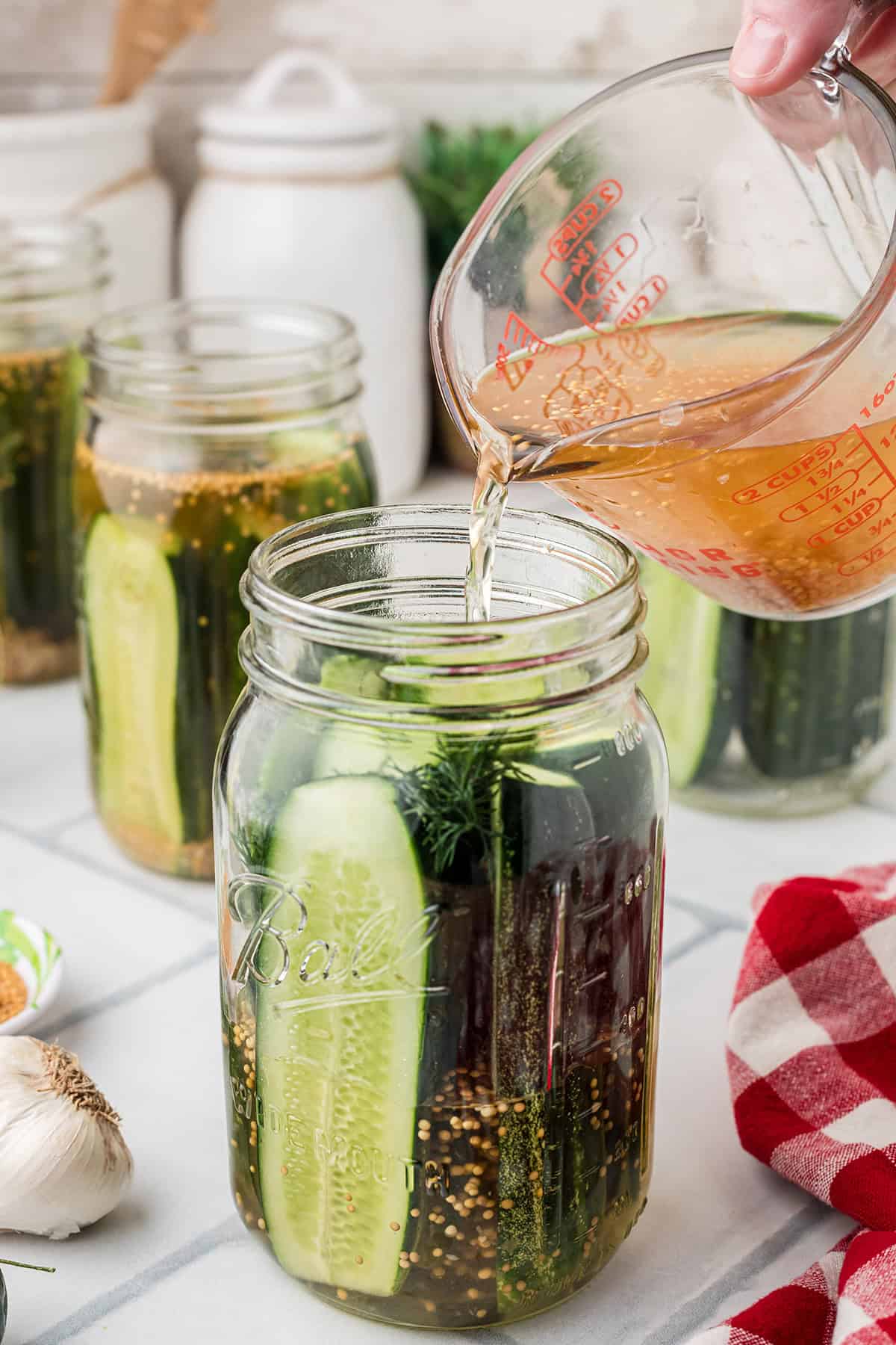 A hand pours pickling brine from a measuring cup into a glass jar filled with cucumbers, dill, and spices, preparing homemade pickles. Other jars and kitchen items are visible in the background.