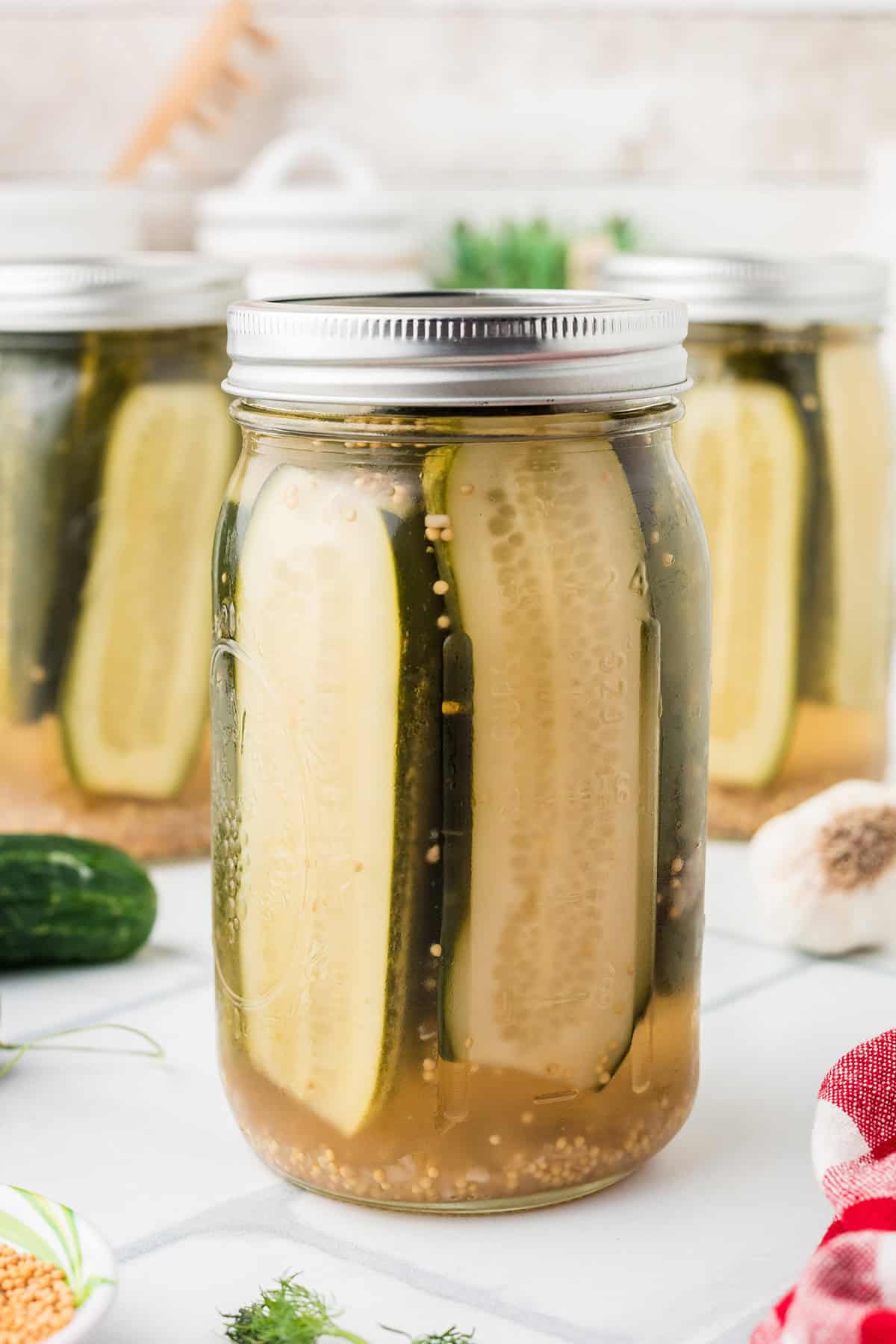 A glass jar filled with pickle spears in brine, with cucumbers, garlic, and herbs visible in the background.