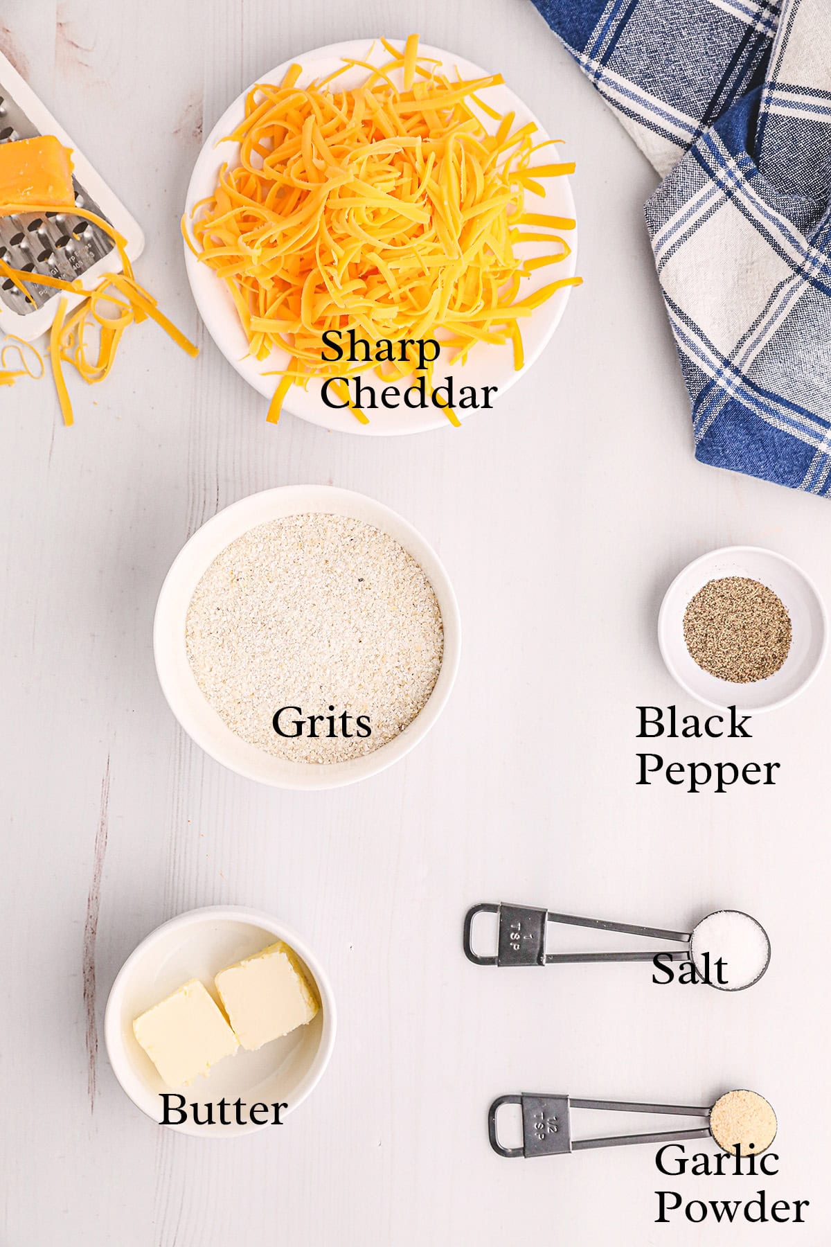 Ingredients for cheese grits: sharp cheddar, grits, black pepper, butter, salt, and garlic powder on a white surface.