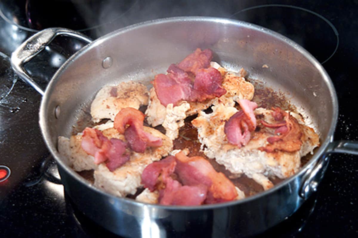 Chicken breasts topped with cooked bacon in a skillet.