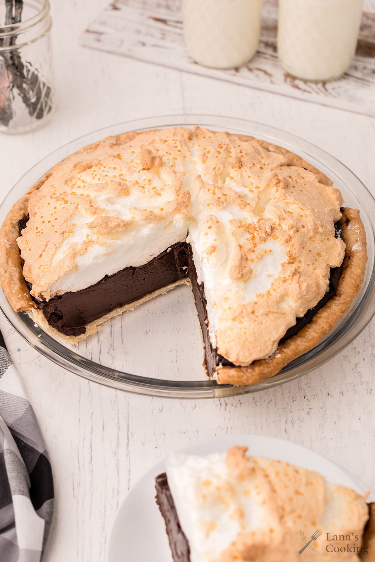 A chocolate meringue pie with a slice missing sits on a white wooden table, next to glasses of milk.