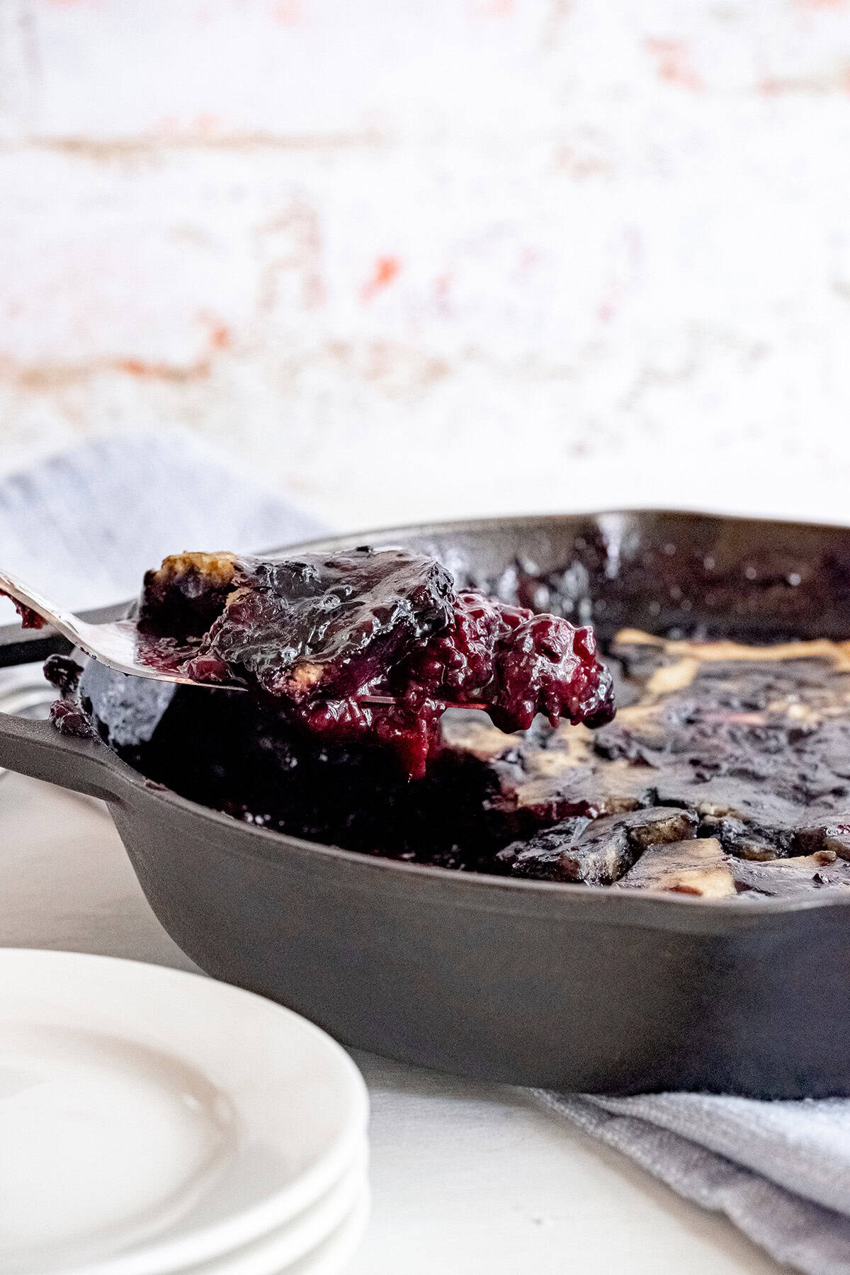 A serving of blackberry cobbler is being lifted from a dark baking dish with a metal spatula; stacked white plates are in the foreground.
