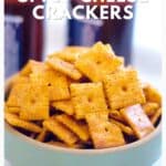 A bowl of square, orange spicy cheese crackers with blurred bottles in the background.