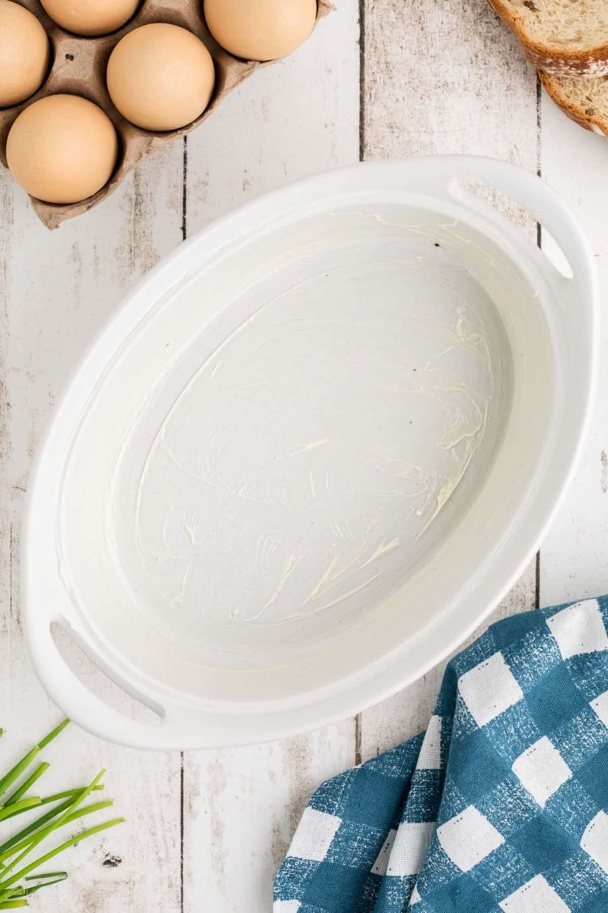 A white oval baking dish with handles sits on a white wooden surface. The dish is greased. Around it are a carton of eggs, a slice of bread, a blue and white checkered towel, and some green herbs.
