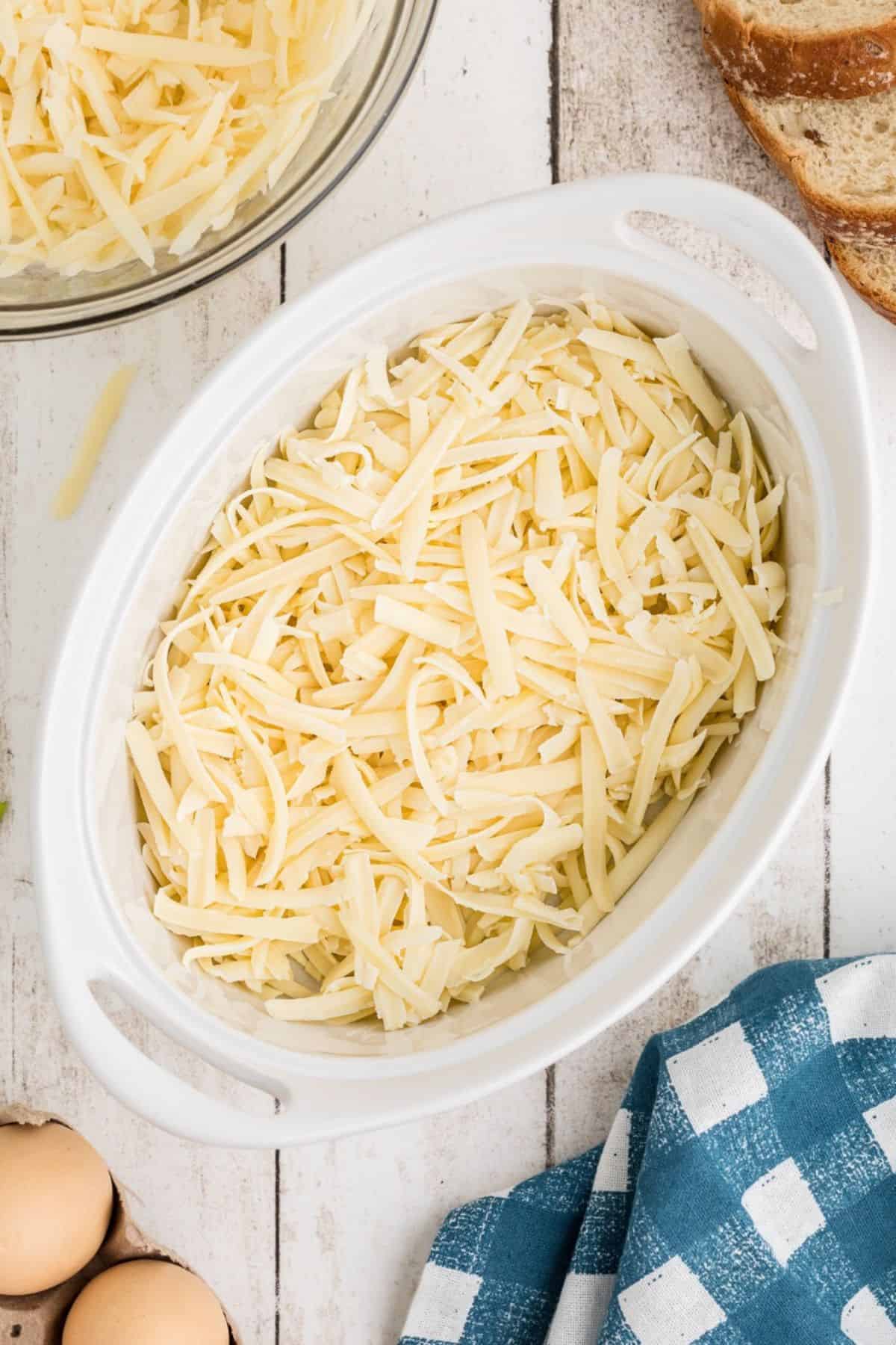 A white oval dish filled with shredded cheese sits on a white wooden surface. Nearby are a blue checkered cloth, two eggs, slices of bread, and a glass bowl with more shredded cheese.