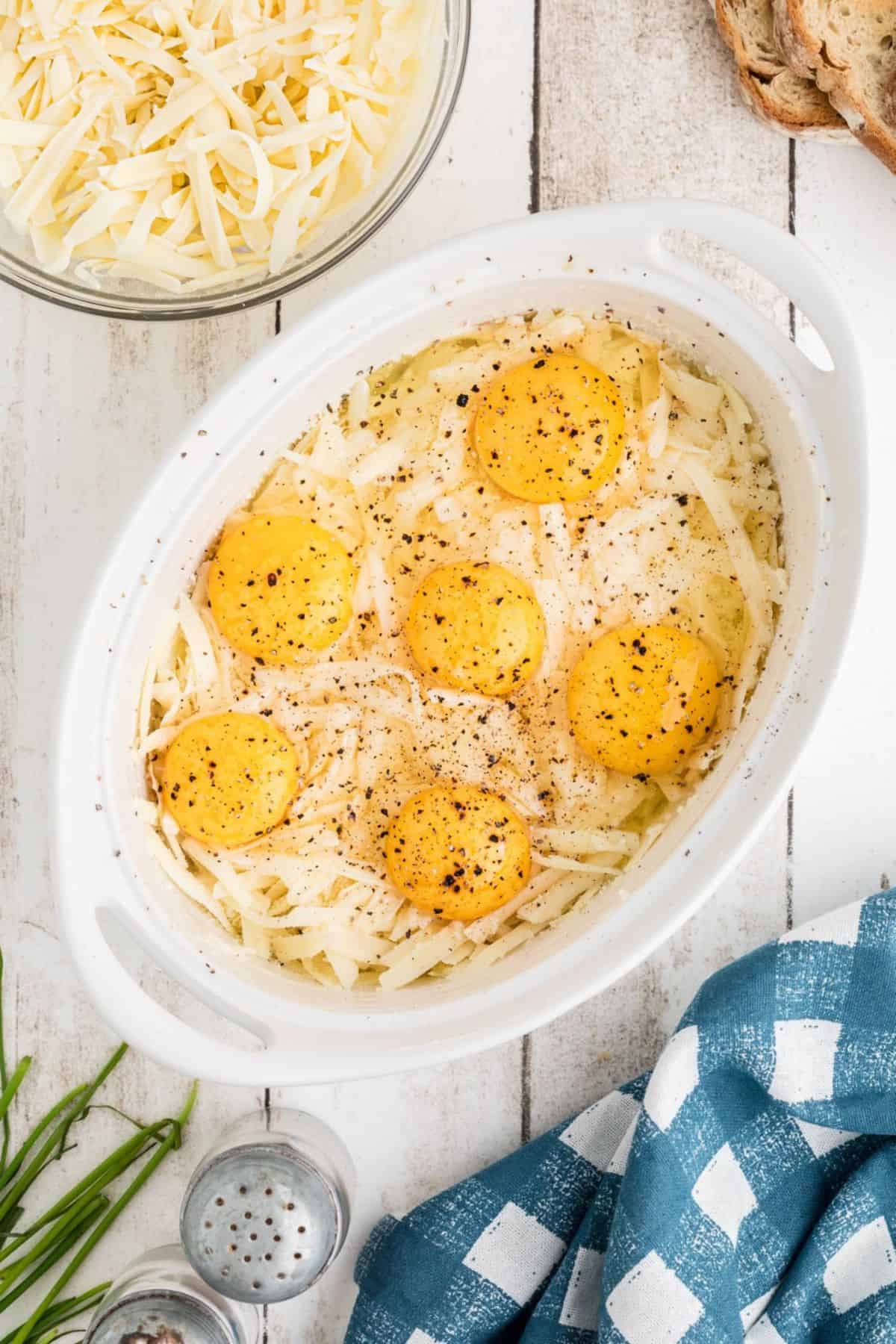 A white oval baking dish filled with shredded cheese, topped with six raw eggs and sprinkled with black pepper, sits on a white wooden surface near a bowl of shredded cheese, bread slices, and a blue checkered towel.