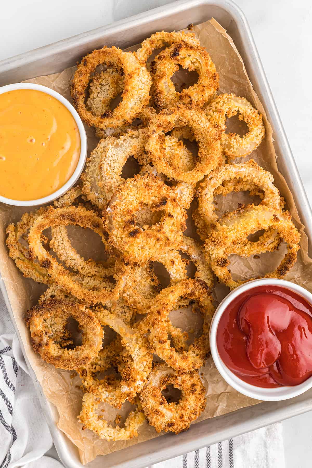 Finished panko onion rings on a baking sheet with a container of ketchup to the side.