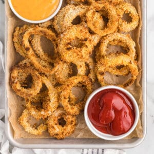 Finished panko onion rings on a baking sheet with a container of ketchup to the side.