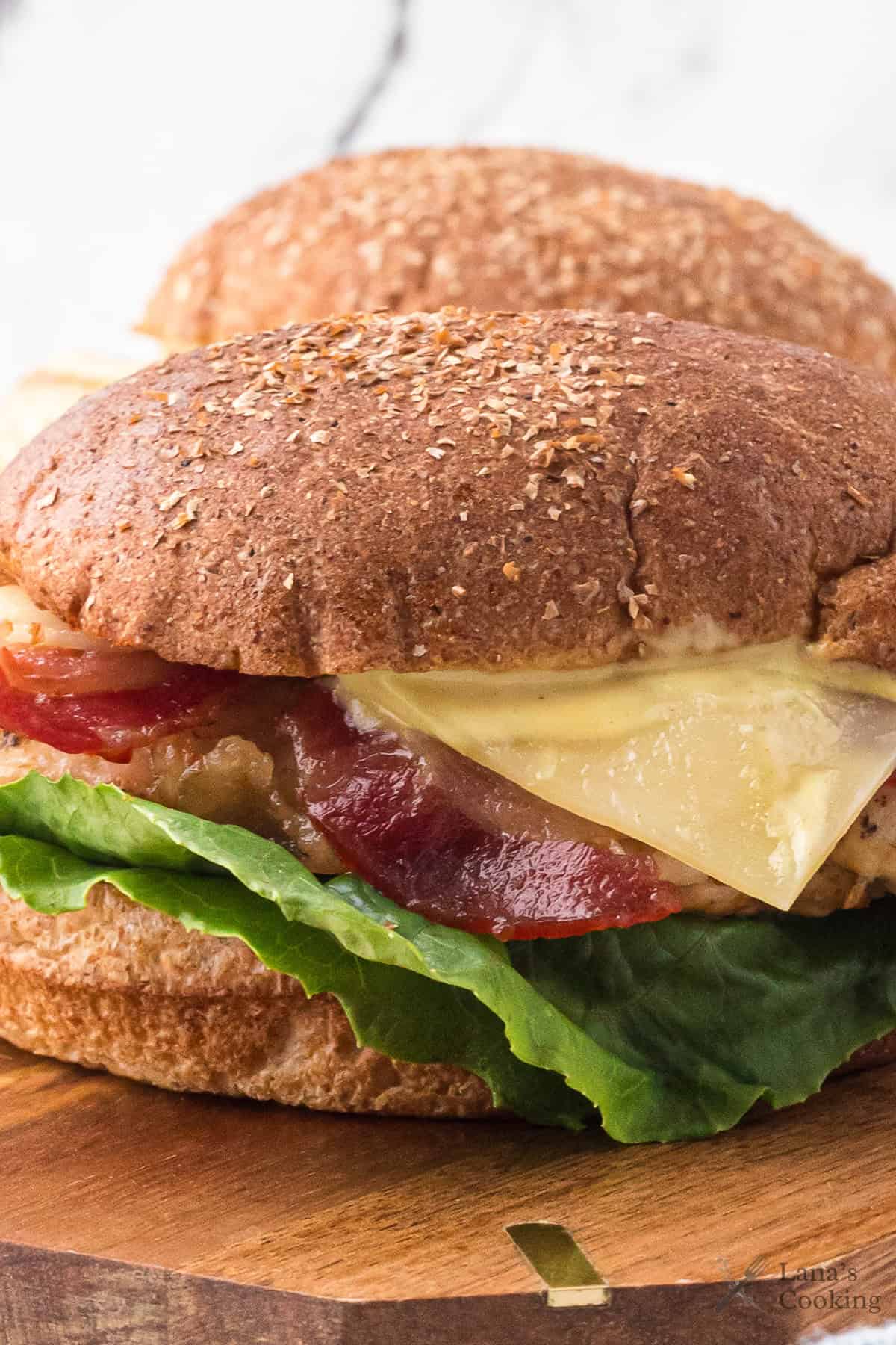 A close-up of a sandwich on a whole wheat bun with lettuce, tomato, melted cheese, and a visible patty, placed on a wooden surface with another sandwich slightly blurred in the background.