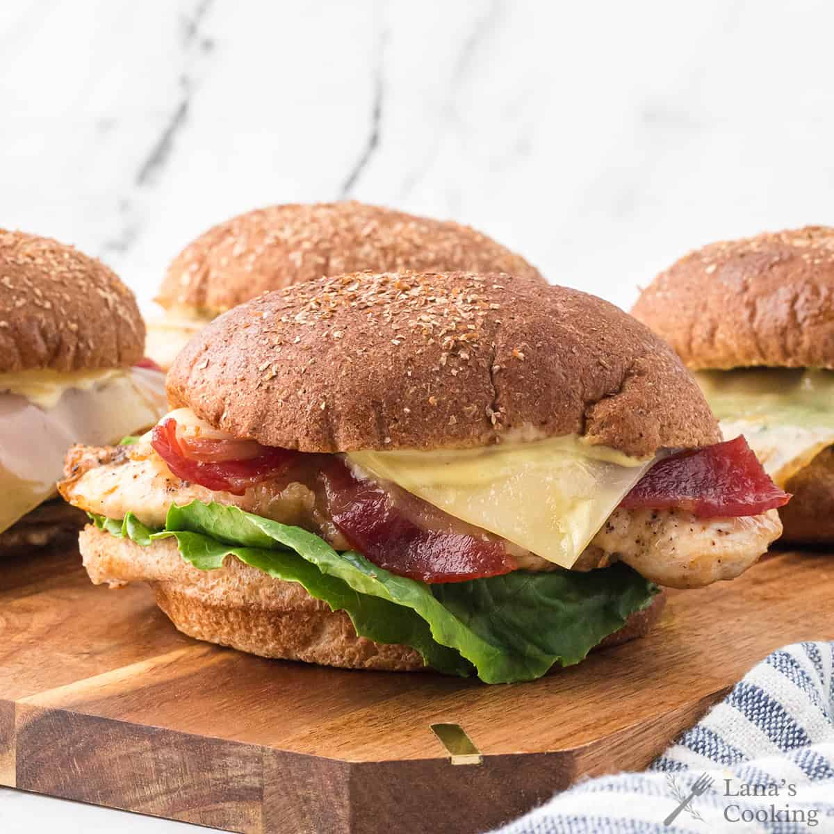Three chicken sandwiches with lettuce, tomato, bacon, and cheese on whole wheat buns are arranged on a wooden board, with a white marble background.