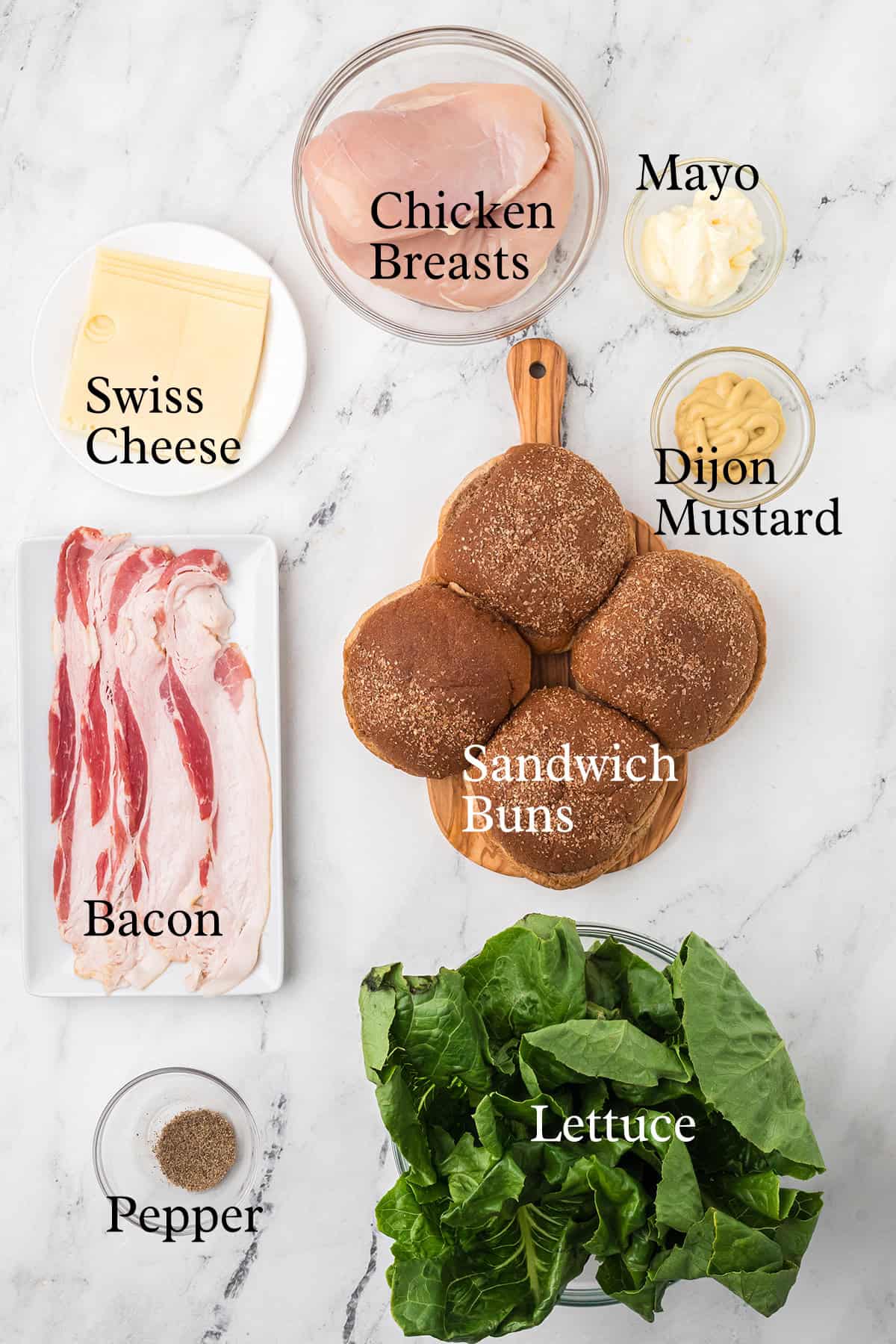 A flat lay of ingredients on a white surface, including chicken breasts, Swiss cheese slices, bacon strips, sandwich buns, lettuce, mayo, Dijon mustard, and ground black pepper. Each item is labeled with text.