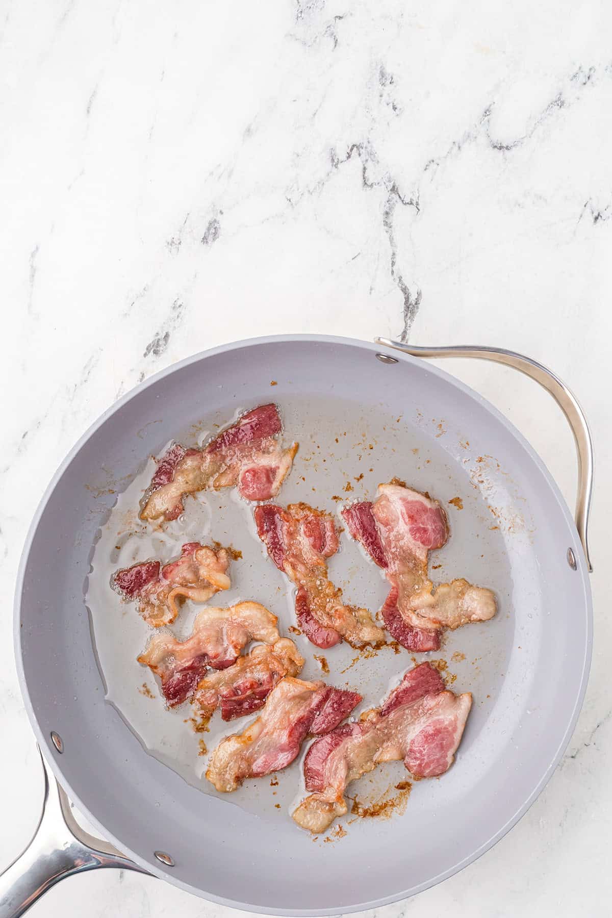 Six strips of bacon cooking in a gray frying pan, with some rendered fat and browned bits visible, on a light marble surface.