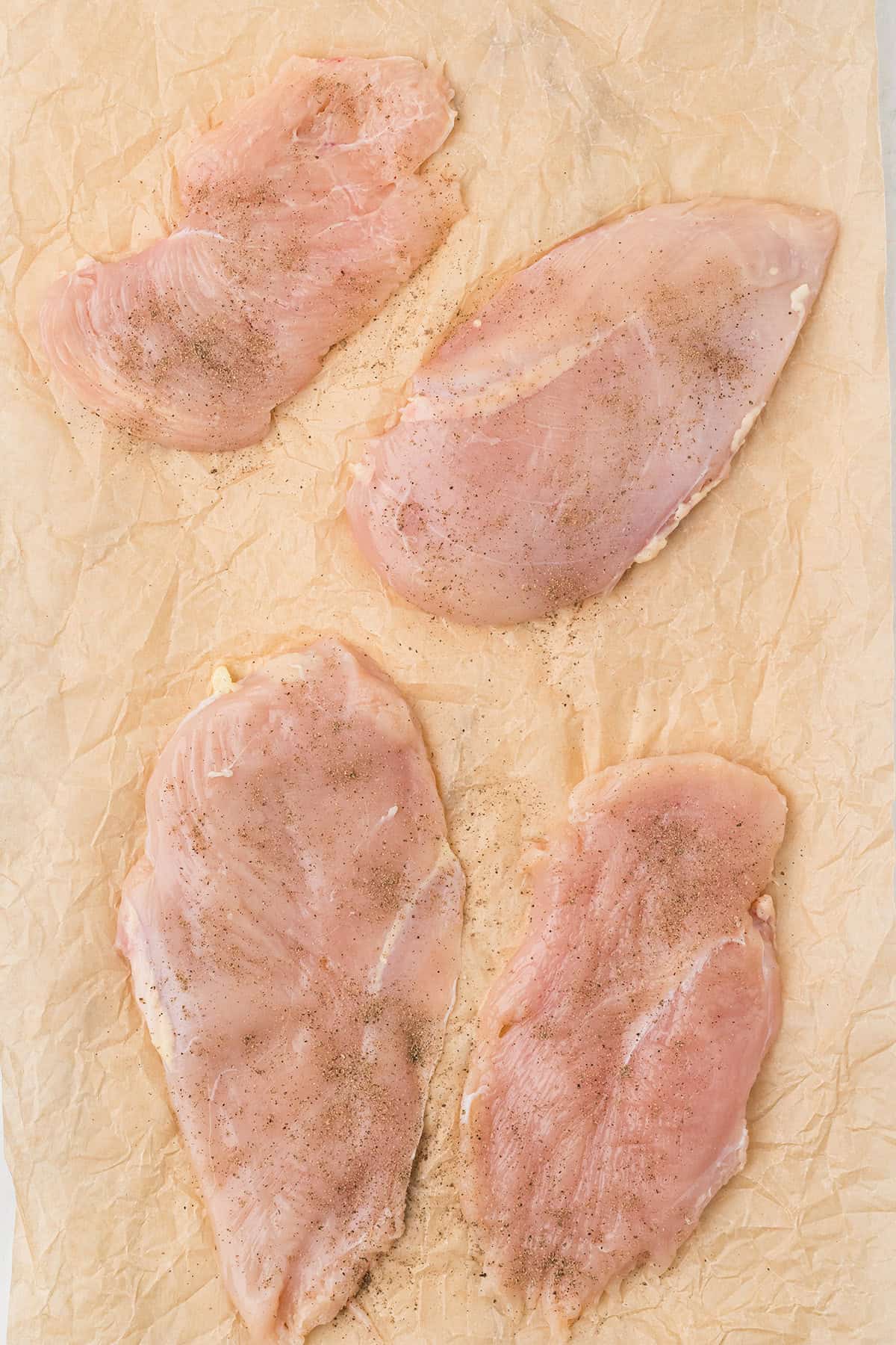 Four raw, thinly sliced chicken breasts are laid out on crumpled parchment paper, lightly seasoned with salt and pepper.