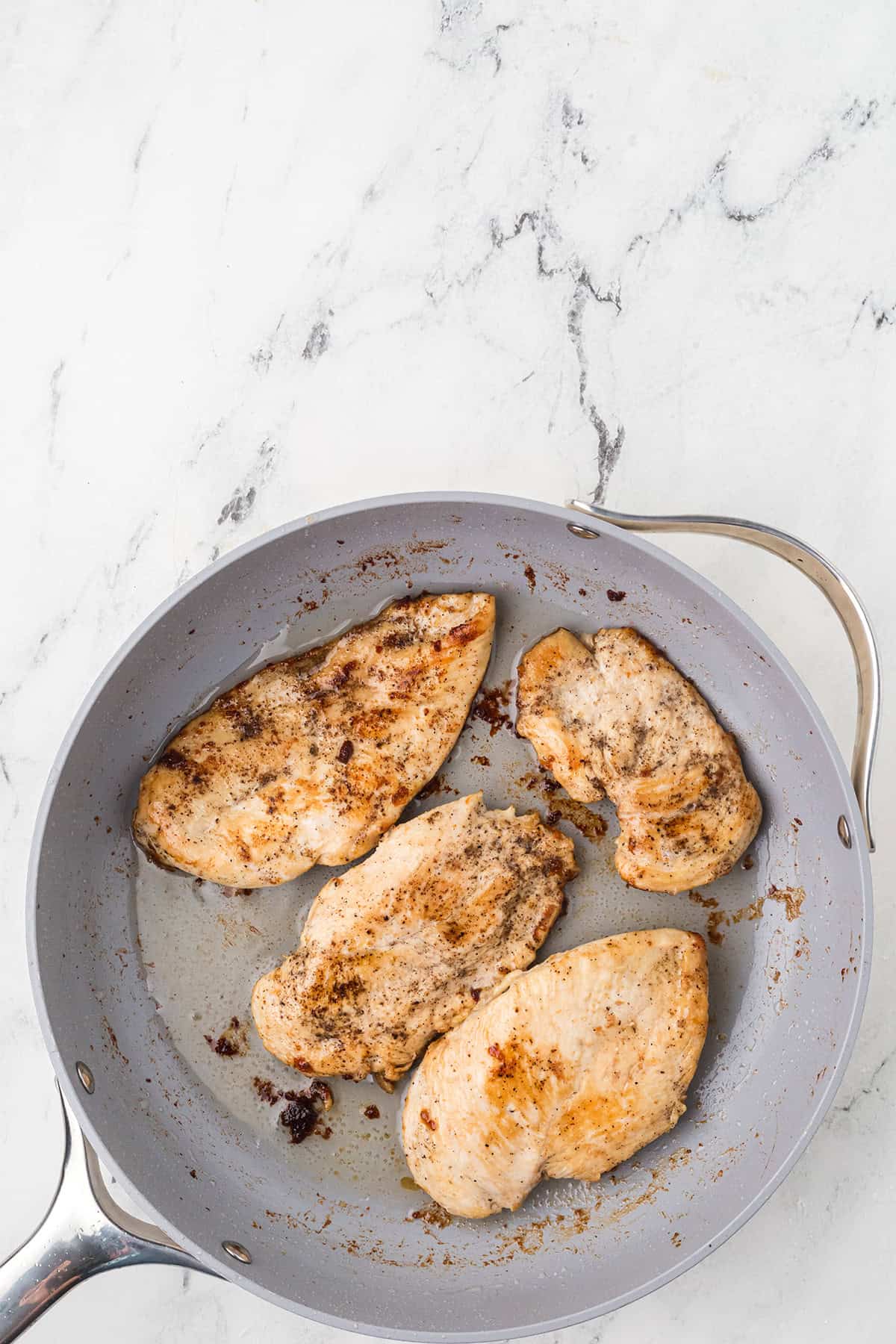 A gray frying pan with four cooked, seasoned chicken breasts on a white marble surface. The chicken pieces have a light brown sear and there are small bits of residue in the pan.