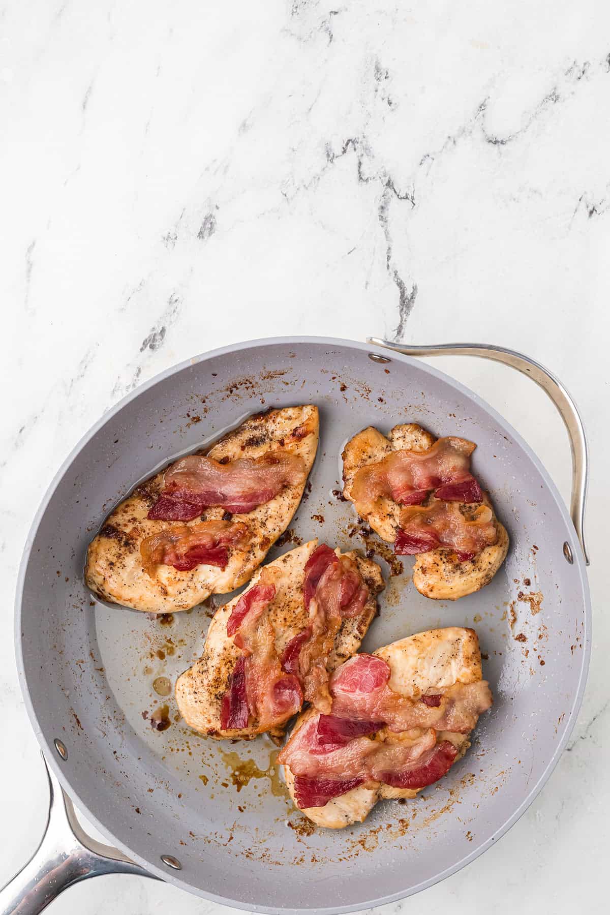 Four pieces of cooked chicken breast topped with bacon strips are in a gray frying pan on a white marble surface. The chicken is browned and the bacon appears crispy. Some oil or juices are visible in the pan.