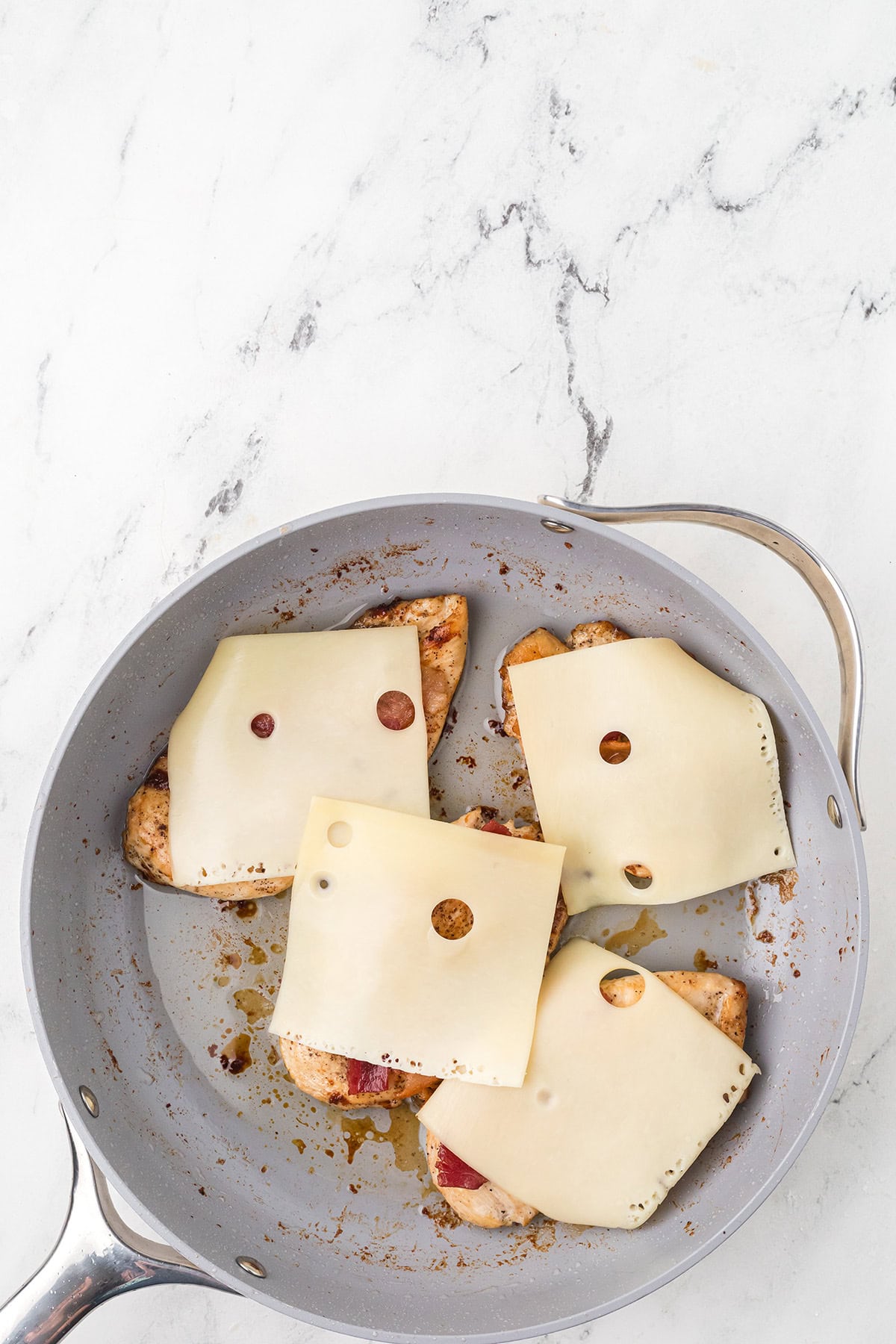 A gray skillet contains four pieces of cooked chicken, each topped with a slice of Swiss cheese. The skillet sits on a white marble surface.
