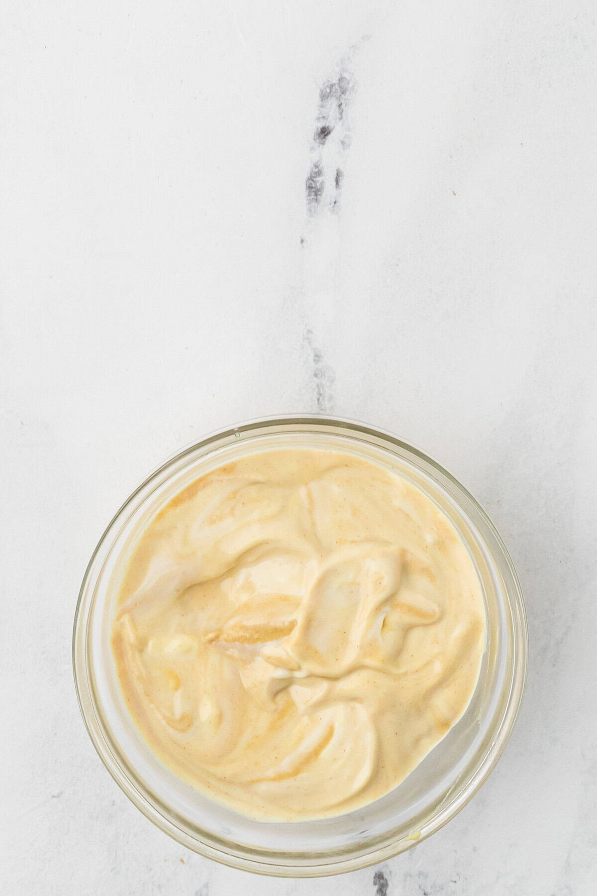 A small glass bowl filled with a creamy, light yellow sauce or dressing sits on a white marble surface.