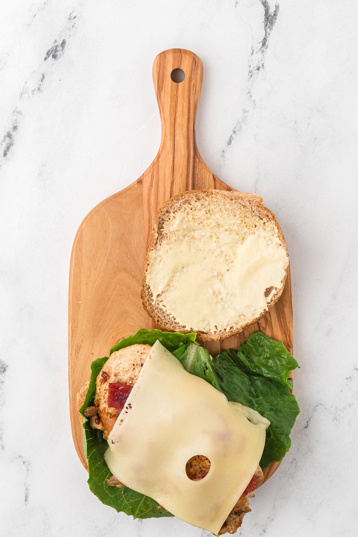 A sandwich with lettuce, a slice of Swiss cheese, and a spread on a sandwich bun sits on a wooden cutting board, with the top half of the bun placed to the side.