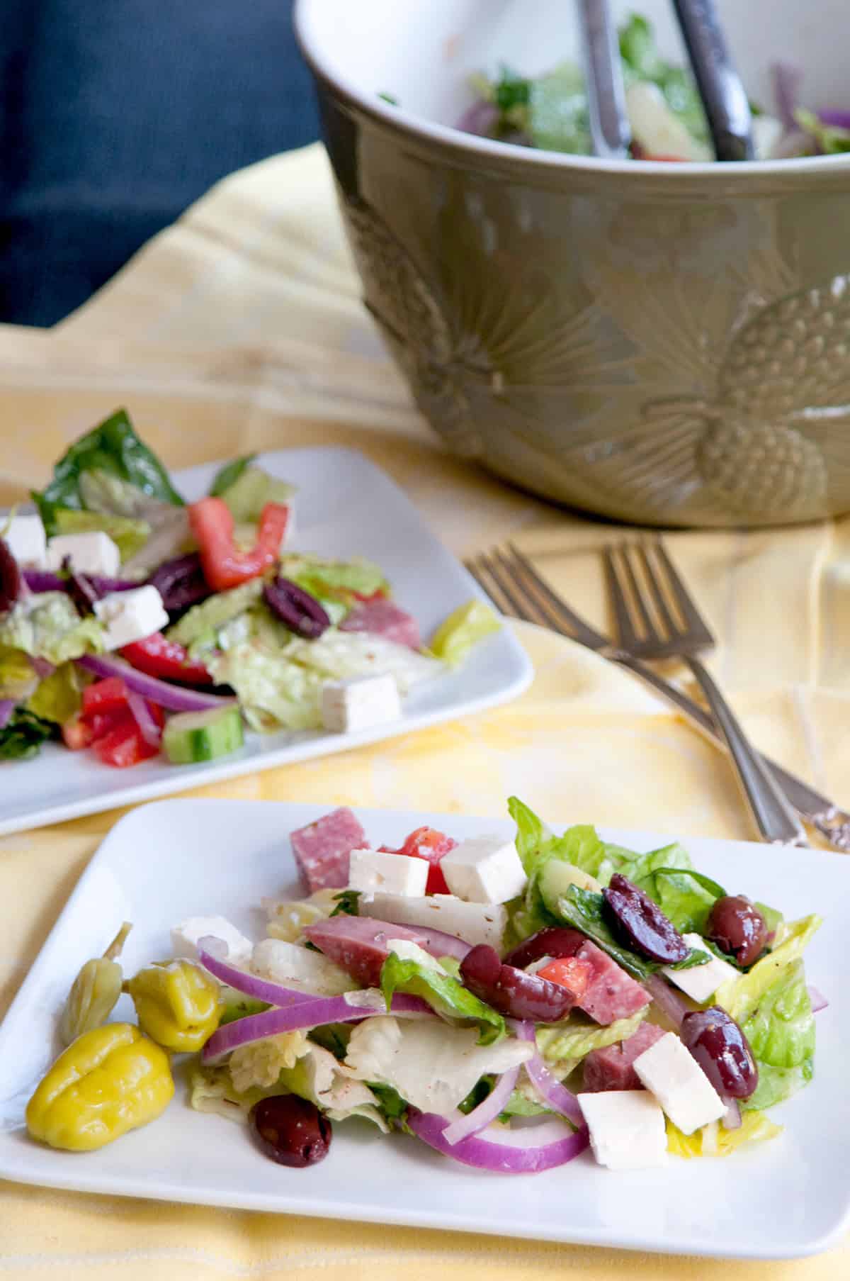 Greek salad with feta, olives, onions, and peppers on plates, with a mixing bowl and forks on a yellow cloth.