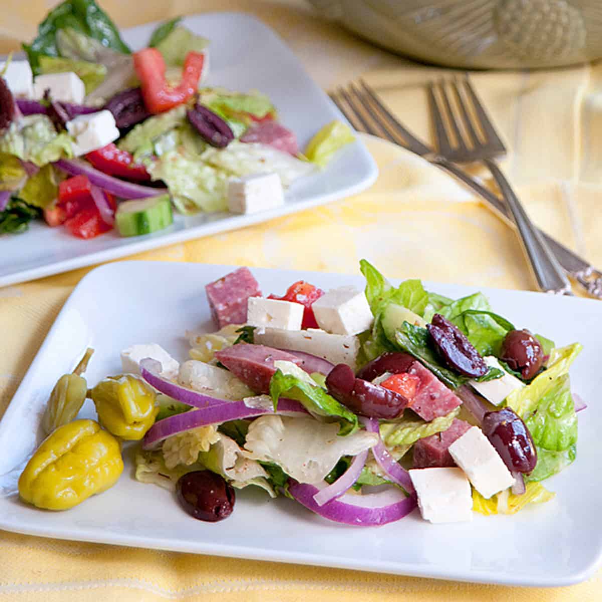 Salad with lettuce, feta, red onion, olives, salami, tomatoes, and pepperoncini on a white plate.